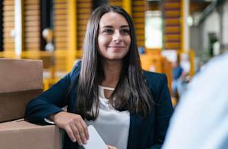 [Featured Image] A project manager in business casual clothing leans against cardboard boxes and talks to their coworker.