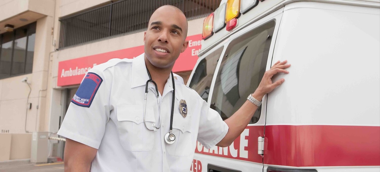 [Feature Image] An EMT in a white button-down shirt with a stethoscope around their neck stands outside the rear of an ambulance.
