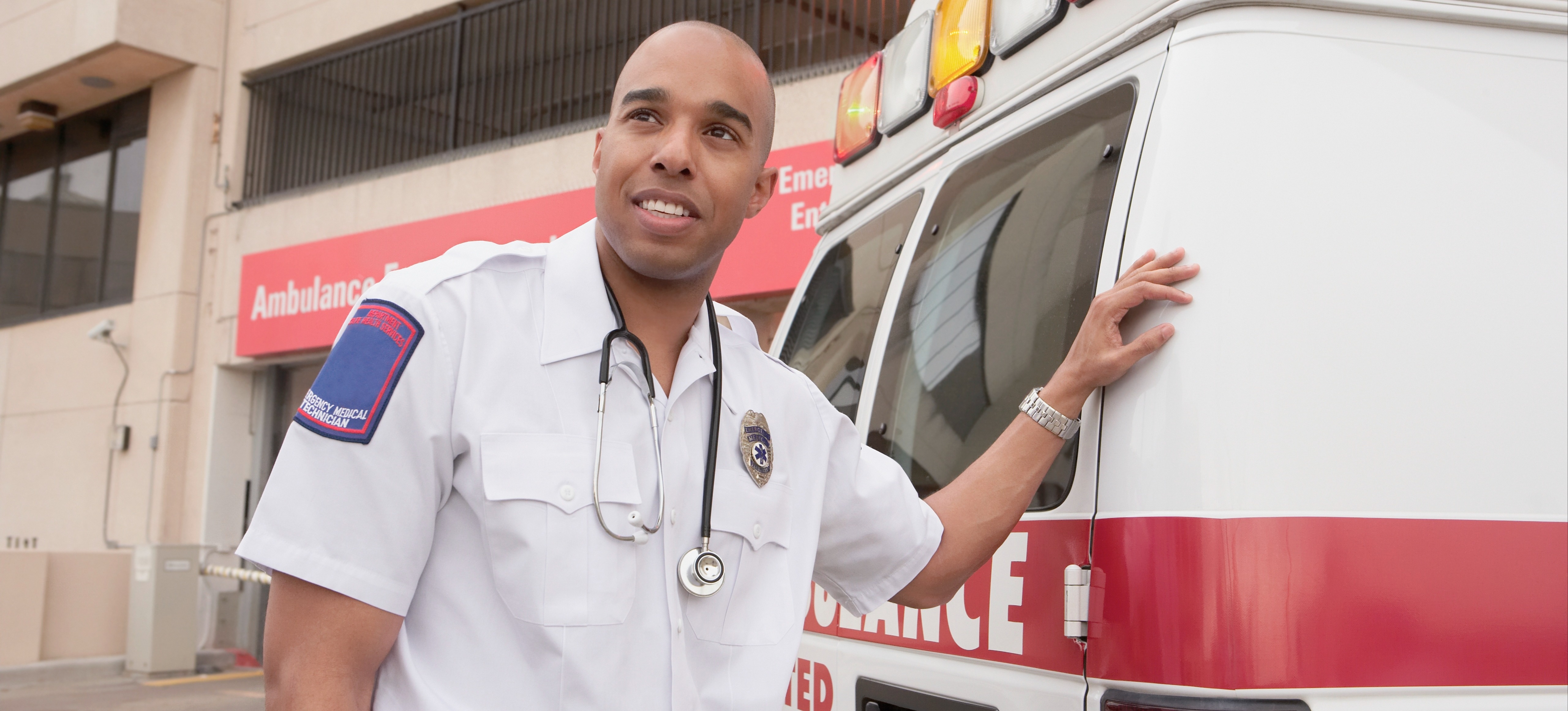 [Feature Image] An EMT in a white button-down shirt with a stethoscope around their neck stands outside the rear of an ambulance.
