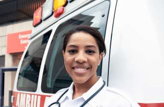 [Featured Image]:  A female paramedic, wearing a white uniform and a stethoscope around her neck, is standing in next to an ambulance, in front of an emergency room entrance. 