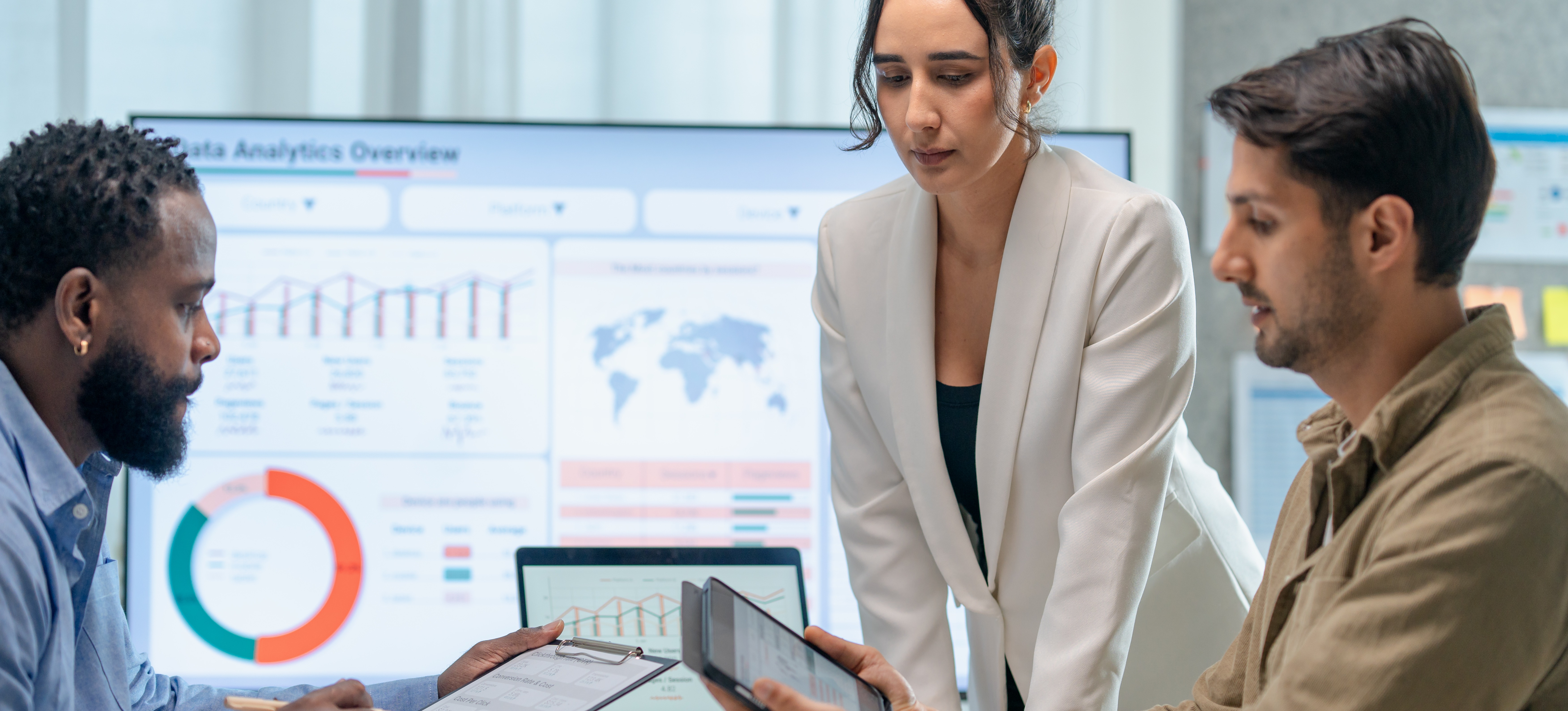 [FEATURED IMAGE] Three marketing professionals work in a conference room looking at marketing analytics on their tablets and computers.
