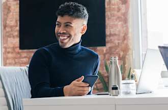 [Featured image] A smiling UX designer works on his phone and laptop in a brightly lit startup office