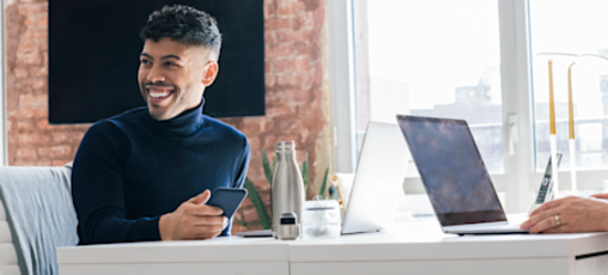 [Featured image] A smiling UX designer works on their phone and laptop in a brightly lit startup office