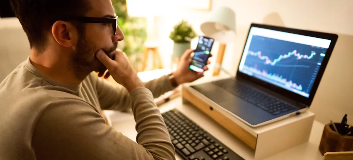 [Featured Image] A person sits in their home office using their phone and laptop to analyze stock market trends using VAE technology.
