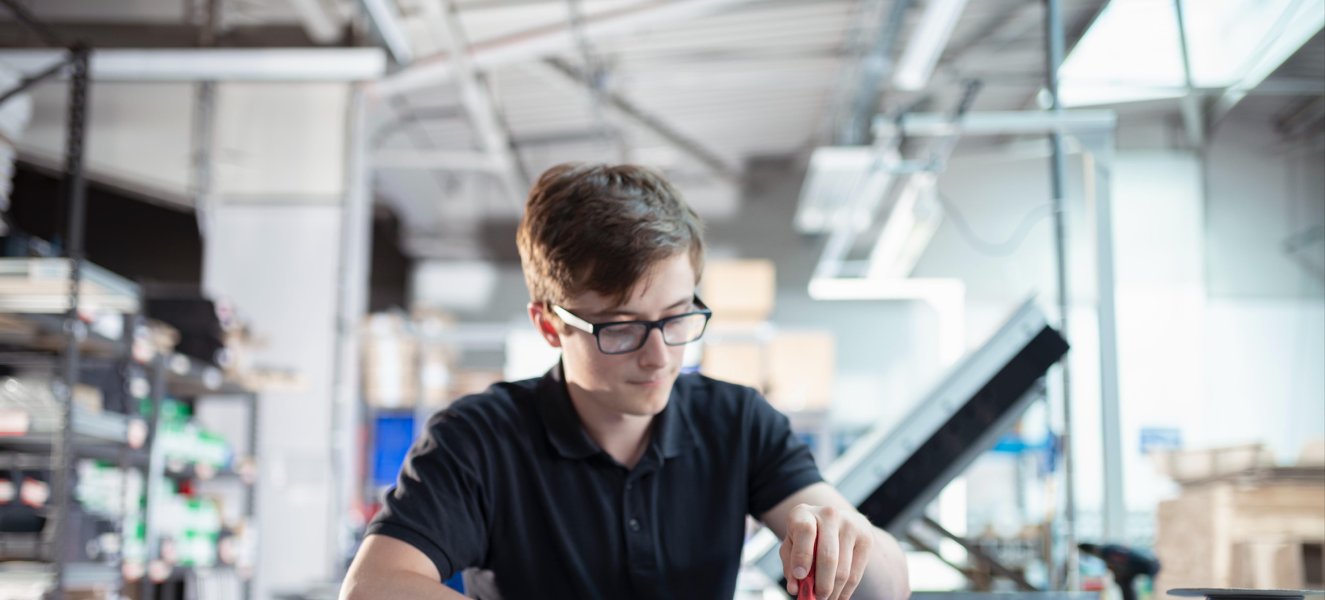 [Featured Image]:  A person, with short brown hair, wearing glasses and a black short-sleeved short, is working on hardware. 