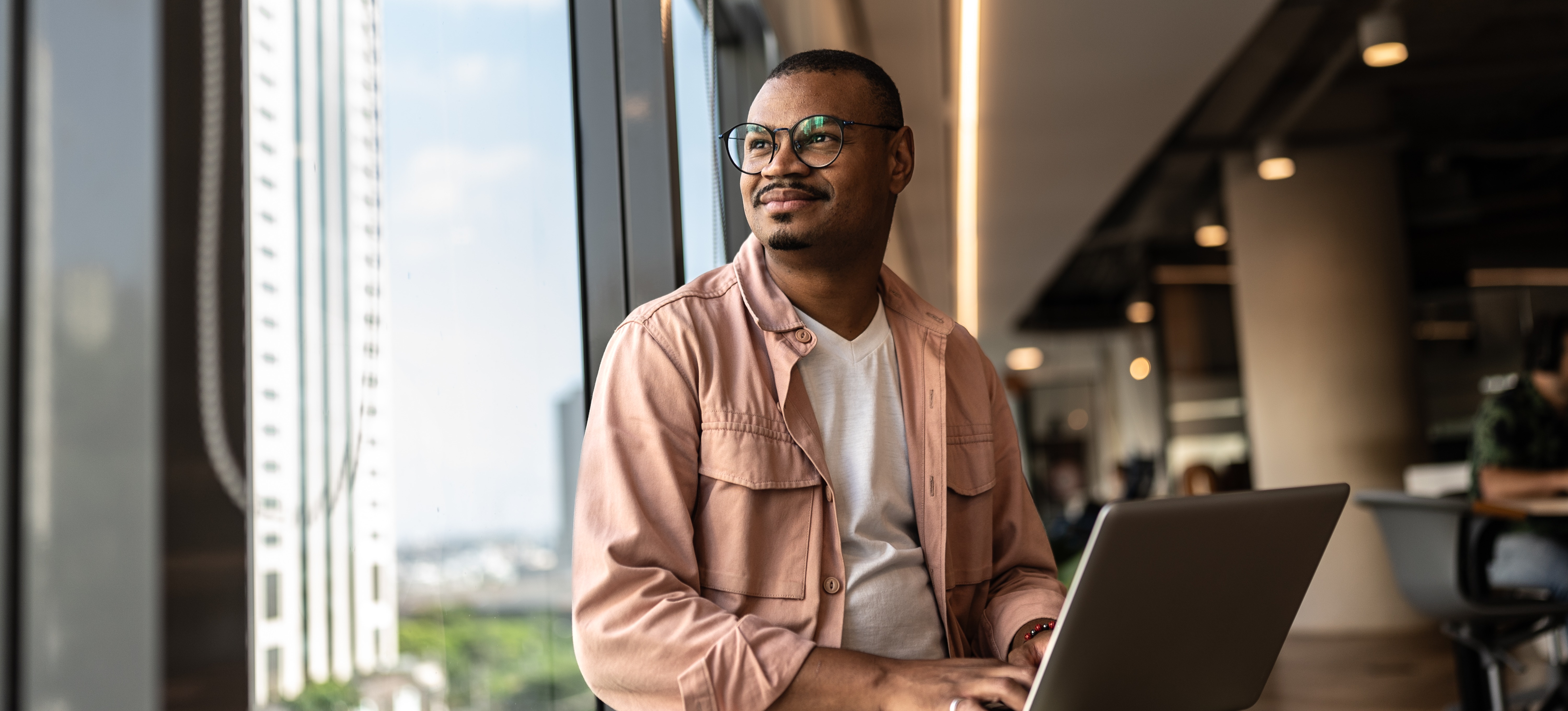[Featured Image] A businessman with hands on his laptop keyboard sits in an office building, thoughtfully looking out the window as he thinks about setting SMART goals.
