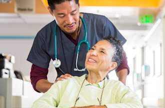 [Featured image] A charge nurse pushes a patient sitting in a wheelchair through a hospital.