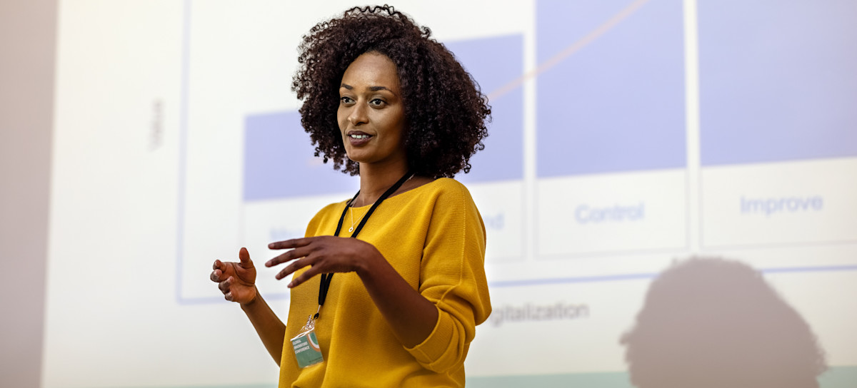 [Featured Image] Data analyst presents in front of a projected screen with essential data visualizations in the background.