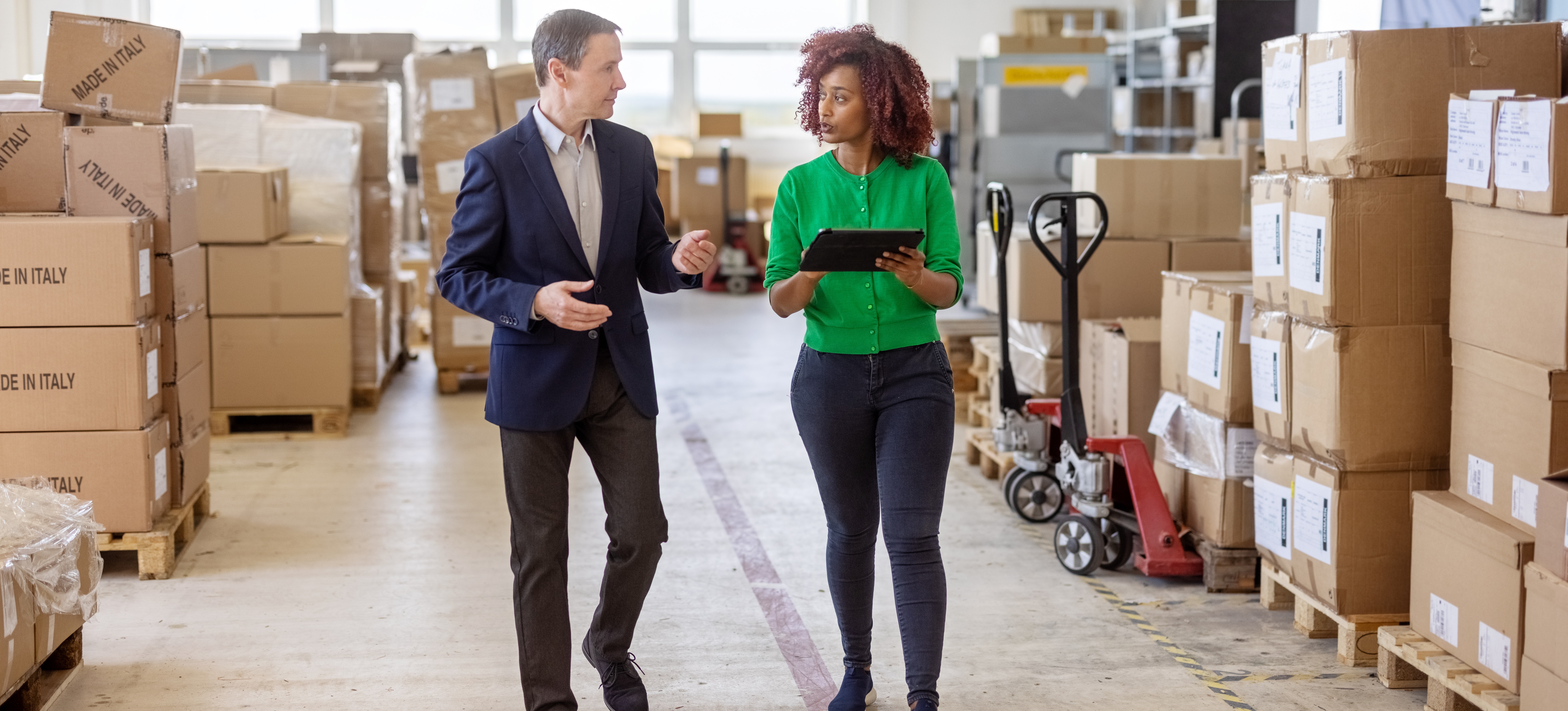 [Feature Image] Two digital supply chain professionals walk through a warehouse, discussing a report they review on their tablet. 
