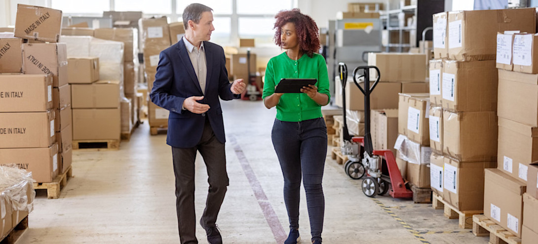 [Feature Image] Two digital supply chain professionals walk through a warehouse, discussing a report they review on their tablet. 

