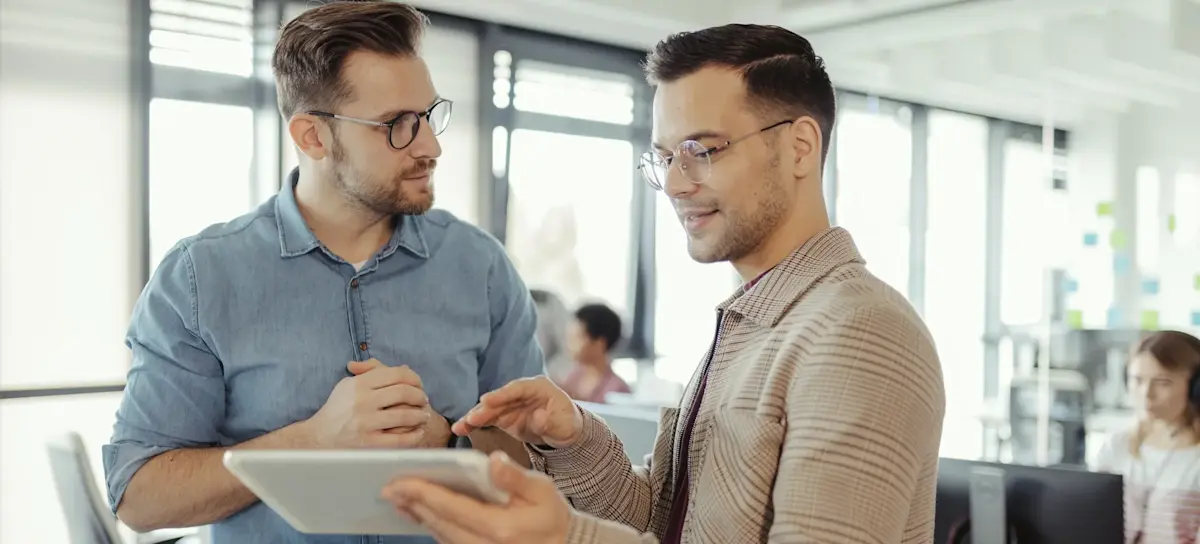 [Featured Image] Two software developers are standing in an open work area consulting GitHub. One developer is holding a tablet while gesturing to it, and the other developer is looking at it intently.
