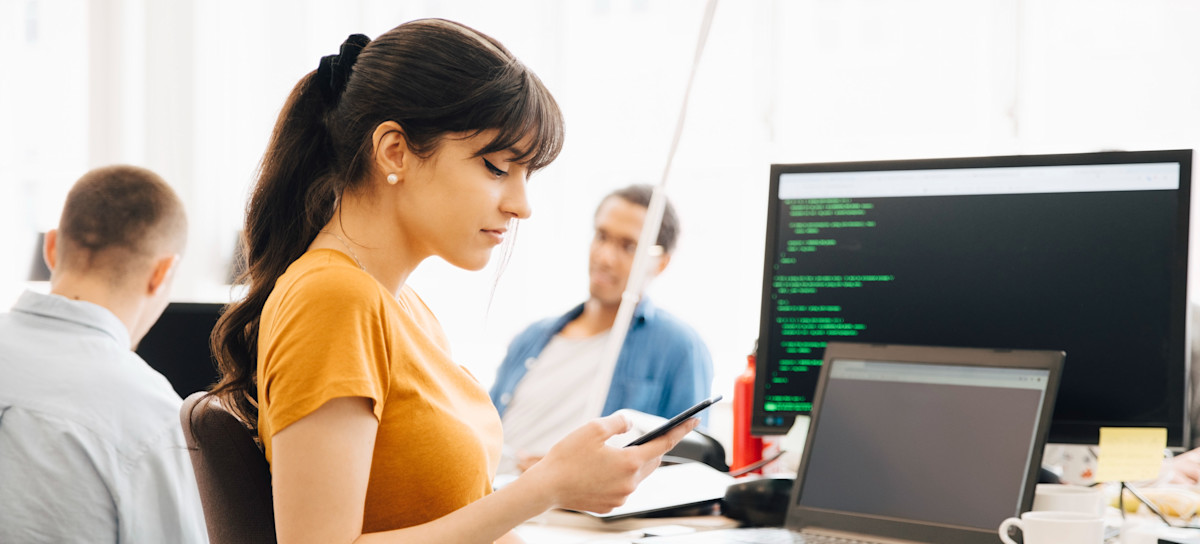 [Feature Image] A learner in an orange short-sleeve shirt takes a break from practicing the front-end languages they’re mastering while sharing a brightly lit study area with two other learners. 
