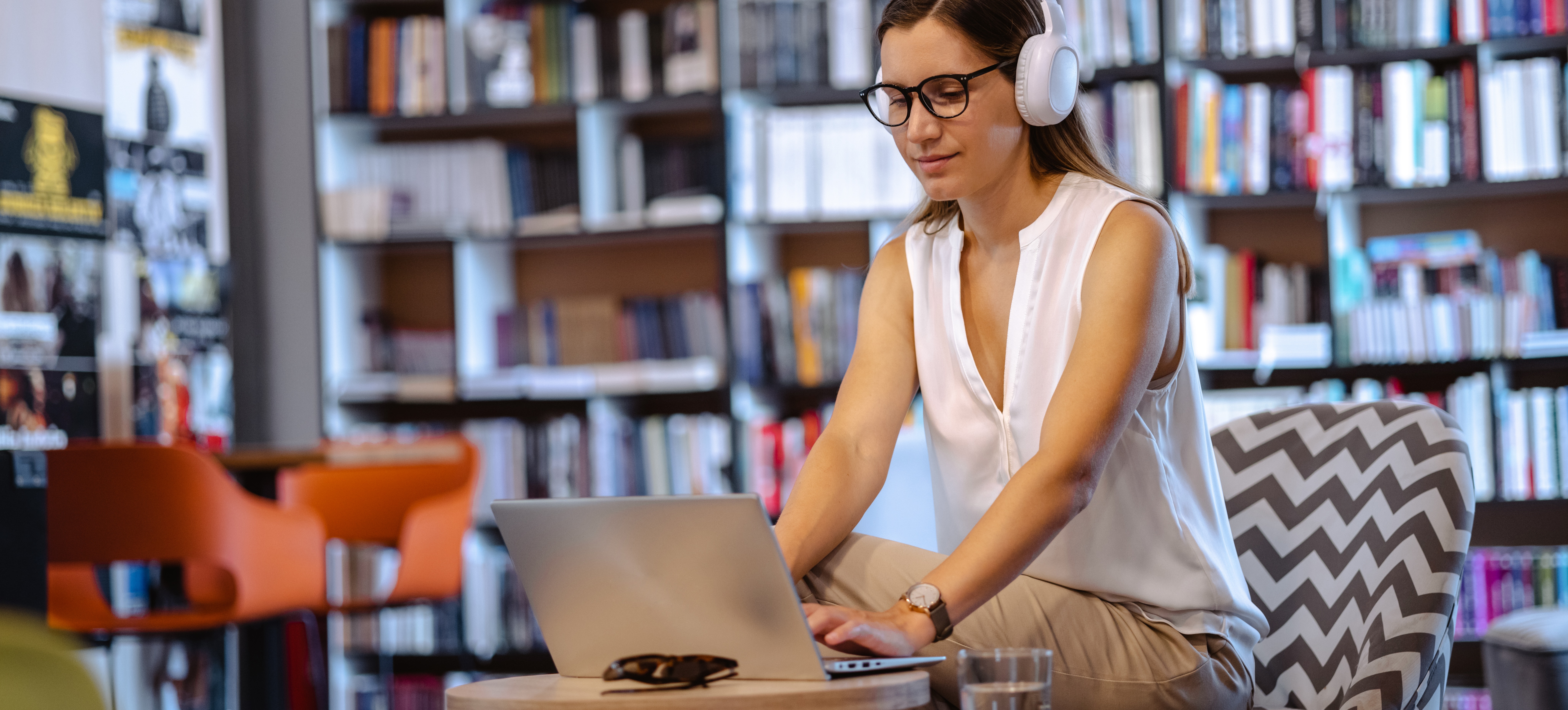 [Featured Image] A college student wearing headphones works on her capstone project on a laptop while sitting in a library.
