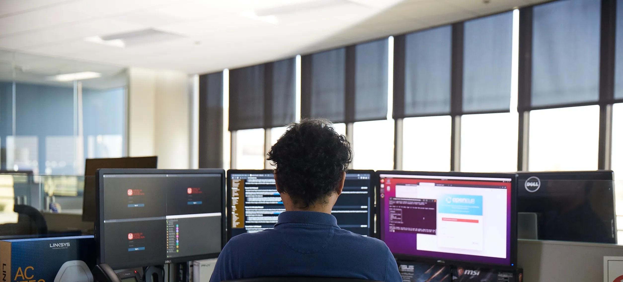 [Featured Image] A person in a dark blue shirt sits facing a trio of computer monitors in a large open-office space. 
