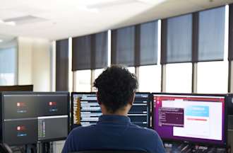 [Featured Image] A person in a dark blue shirt sits facing a trio of computer monitors in a large open-office space. 