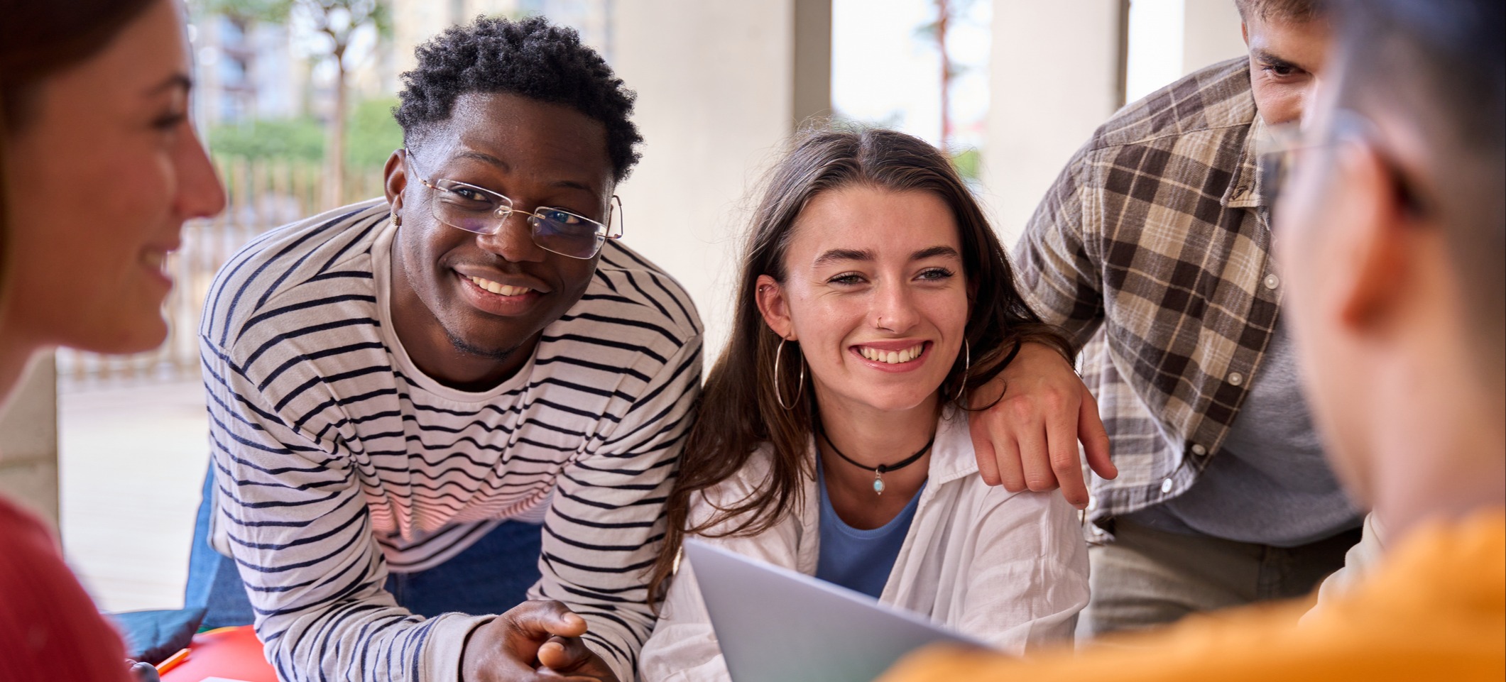[Featured Image] A group of happy students deciding between trade school vs college discuss the pros and cons in their classroom.