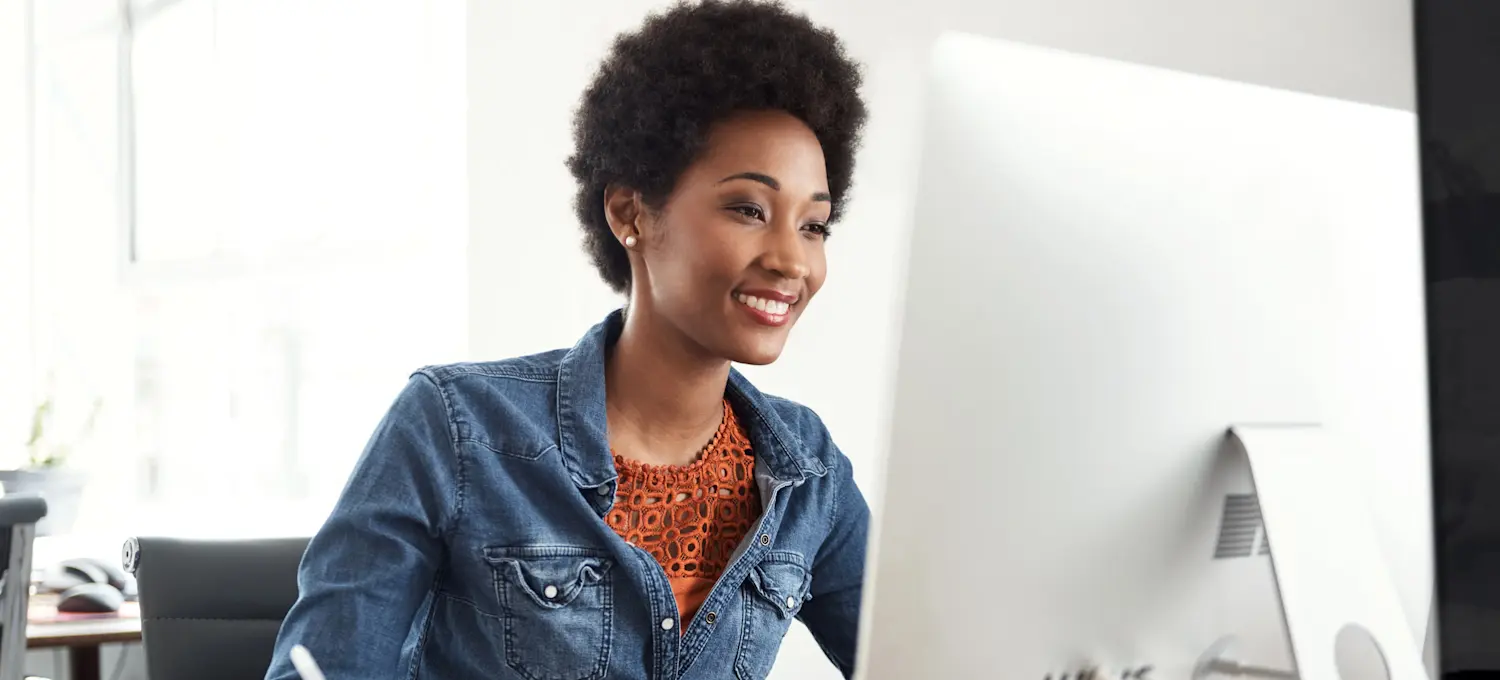 [Featured Image] A smiling professional dressed in denim using an email marketing strategy holds a pen above a journal and looks at their computer screen.