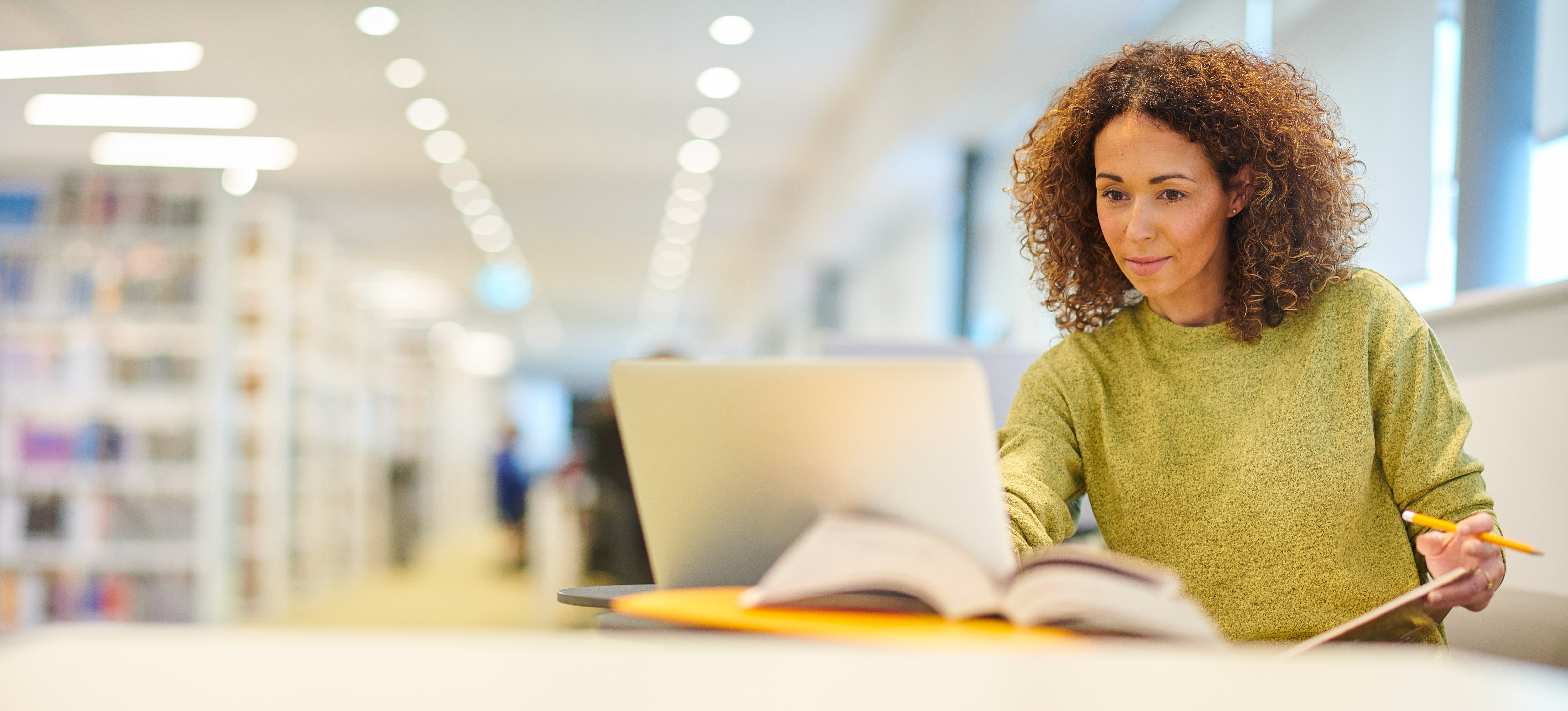 [Featured Image] A student sits in a library with a laptop, books, paper, and pencil, using their research skills to write an essay.
