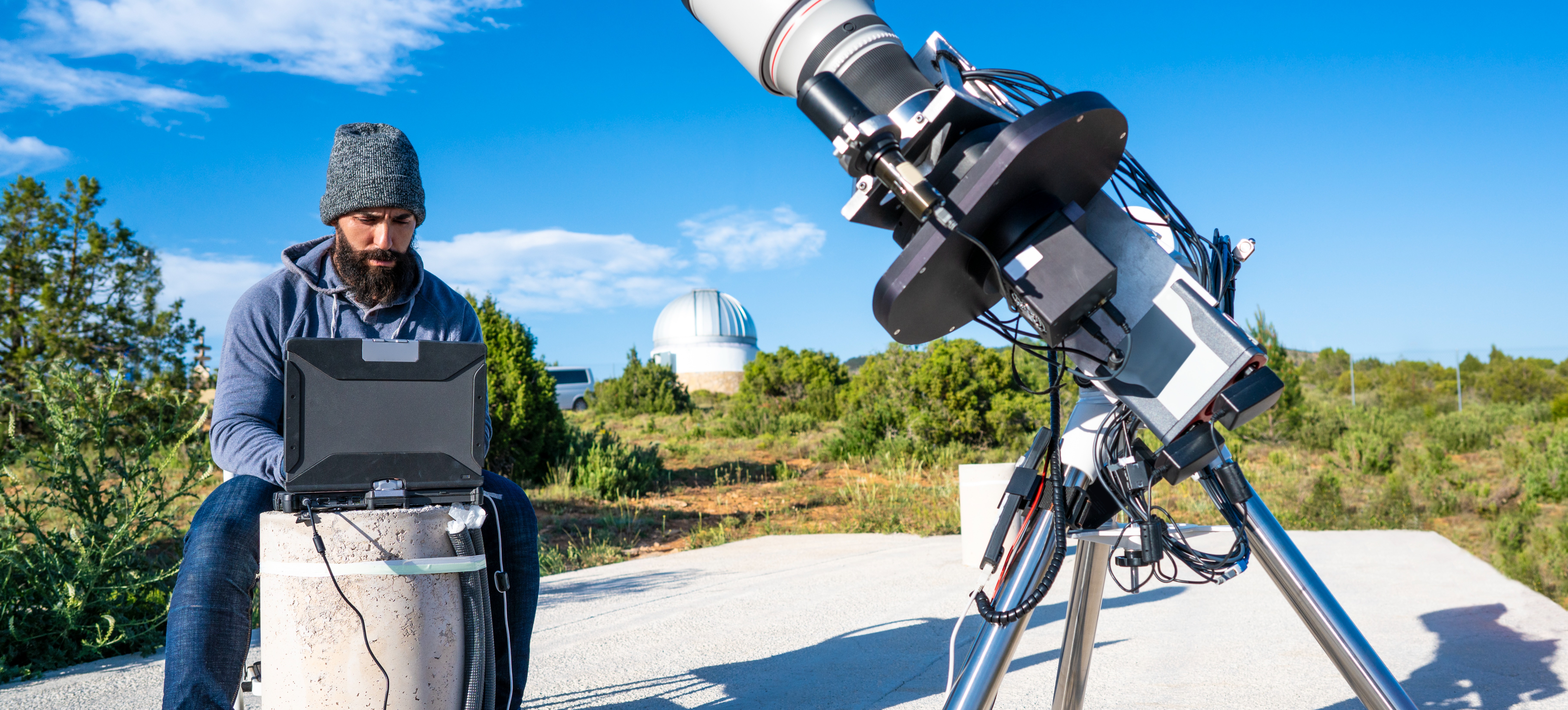[Featured Image] As part of their NASA job, an astronomer connects a telescope to a computer while on a cement platform in the middle of a grassy field.