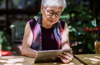 An older woman sits outside at a wooden table reading a book on her tablet device.