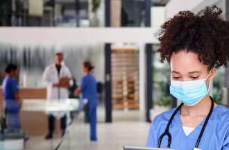 [Featured Image]: A woman with curly hair, wearing blue scrubs, a mask and a stethoscope around her neck is reading a chart. Doctors and nurses are in the background.