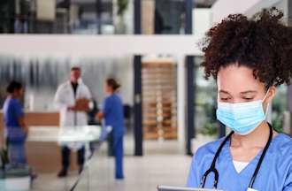 [Featured Image]: A woman with curly hair, wearing blue scrubs, a mask and a stethoscope around her neck is reading a chart. Doctors and nurses are in the background.