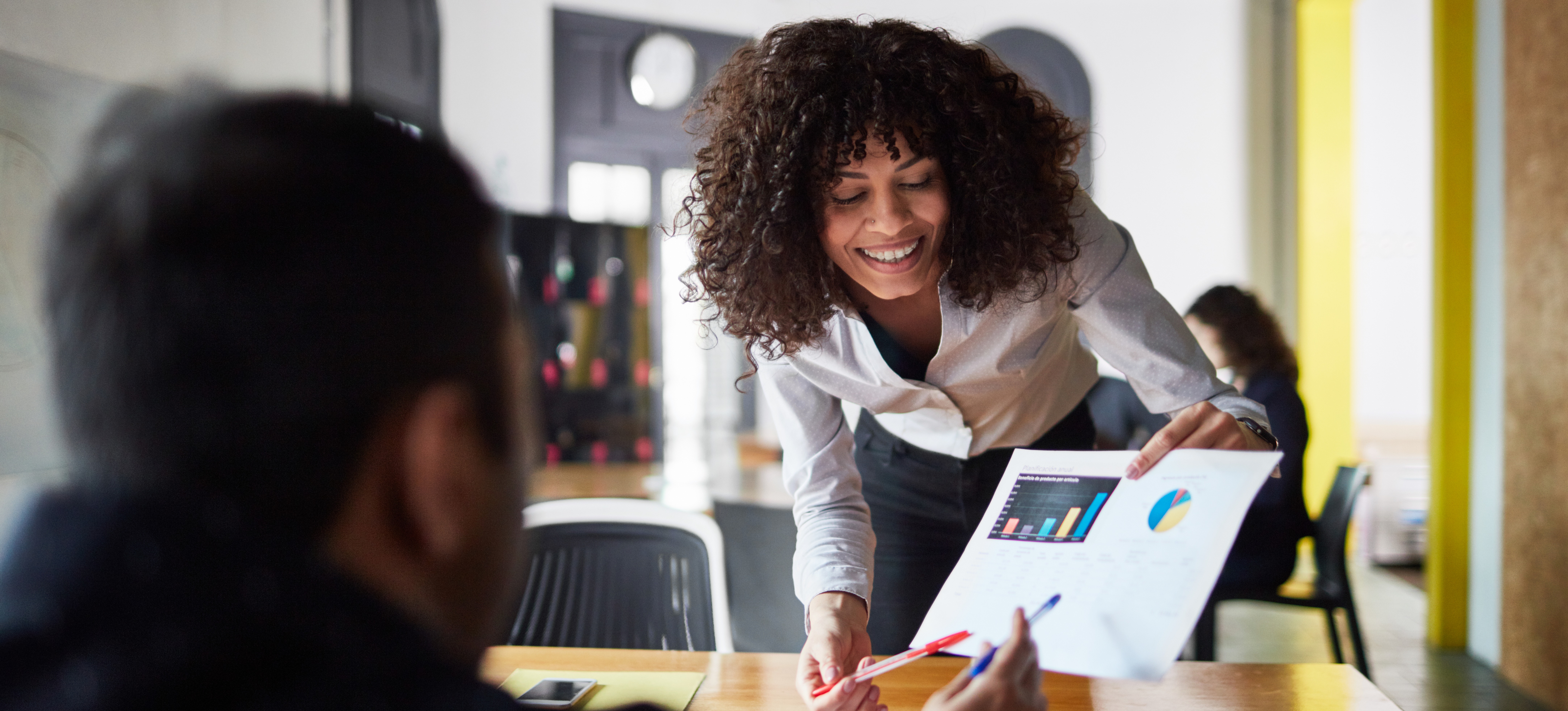 [Featured image] A person in a white blouse leans across a table to show a colleague a printout with data visualizations to explain what data science is. 