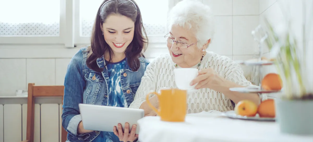 [Featured Image] A young person sits in a kitchen with a tablet and volunteers with a senior citizen to help improve their service orientation skills.
