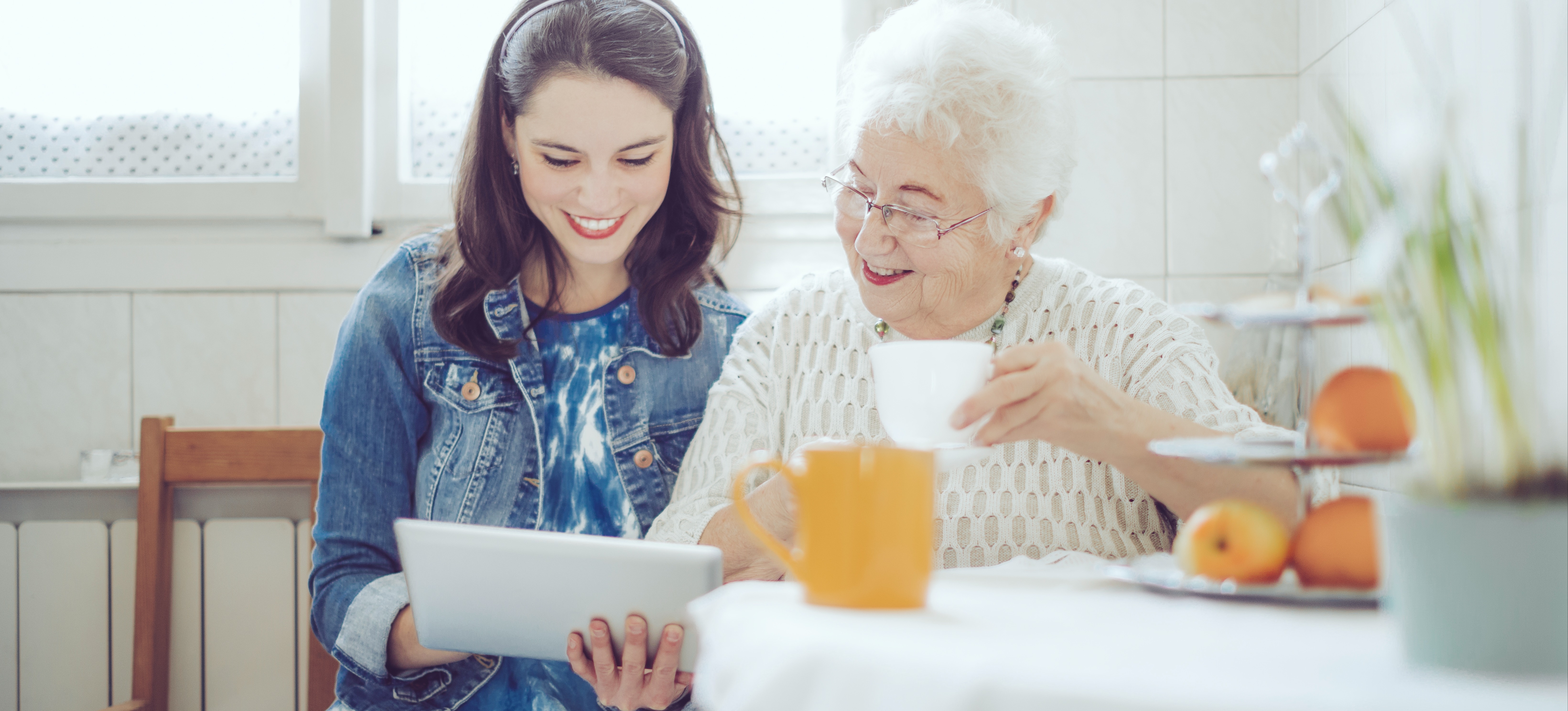 [Featured Image] A young person sits in a kitchen with a tablet and volunteers with a senior citizen to help improve their service orientation skills.
