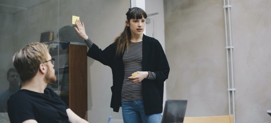 [Imagen destacada] Dos compañeras de trabajo vestidas con blusas negras trabajan en un proyecto de diseño de usuario (UX) con un portátil en una sala de conferencias con una pared de cristal.
