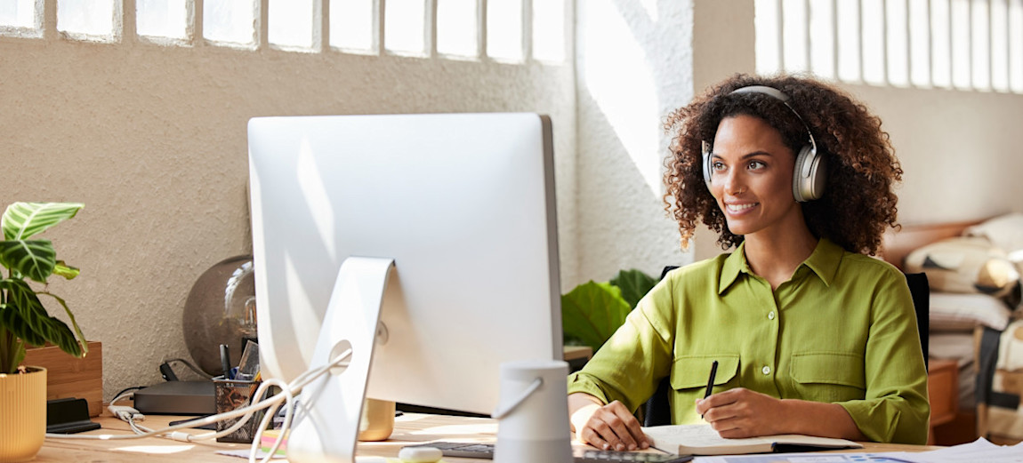 [Featured Image] A learner wearing silver headphones sits at a light wood desk in a room with sunlight streaming through the windows, smiling as they learn about data mart vs. data warehouse.

