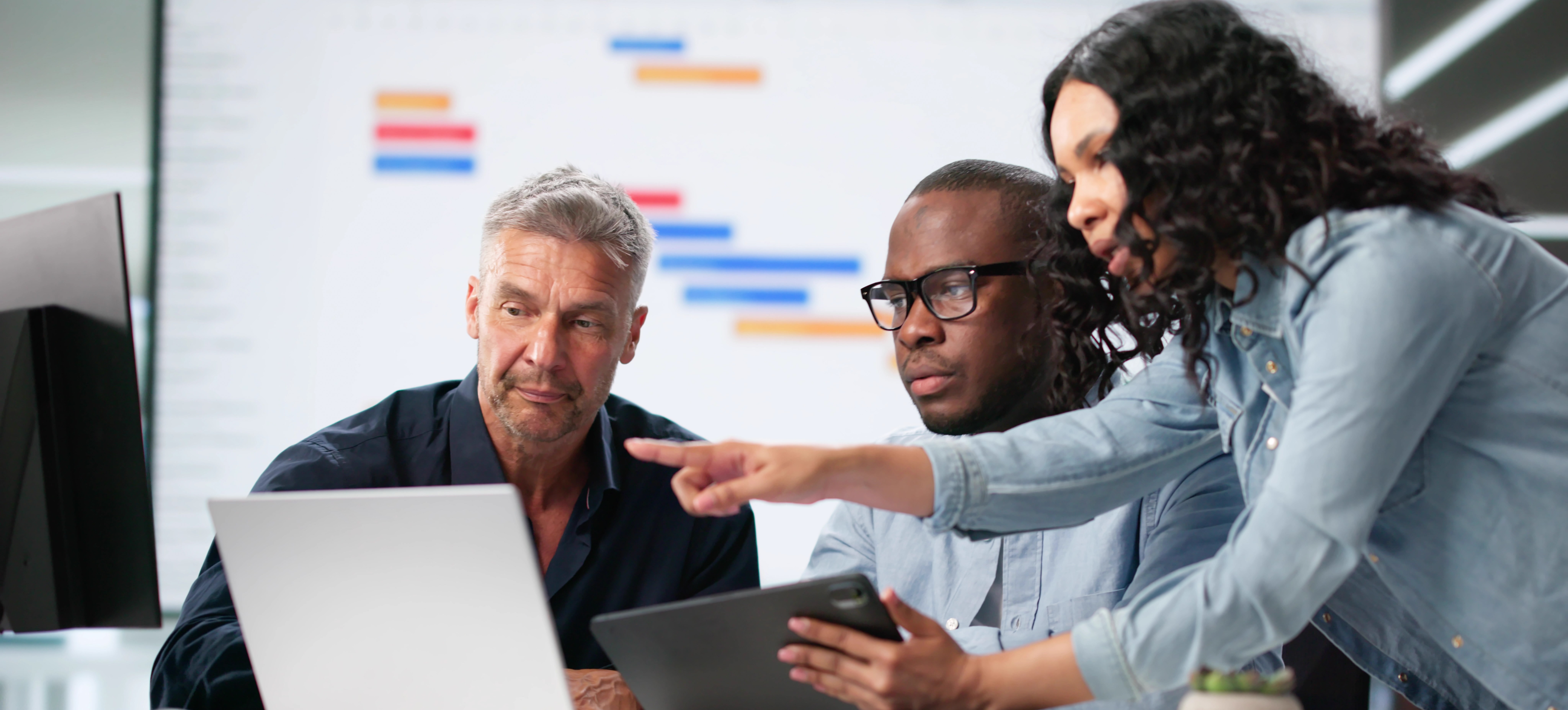 [Featured Image] Three colleagues discuss how to automate a process at work while looking at a computer screen and a tablet.
