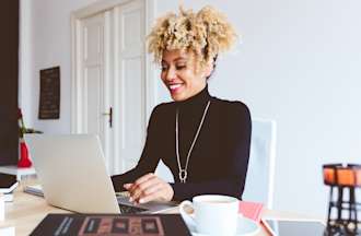 [Featured image] A smiling woman in a black shirt and necklace sits at her laptop and works on her UX portfolio in a brightly lit office.