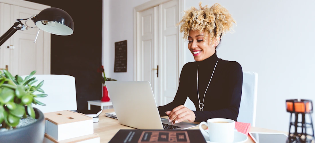 [Featured image] A smiling person in a black shirt and necklace sits at their laptop and works on their UX portfolio in a brightly lit office.
