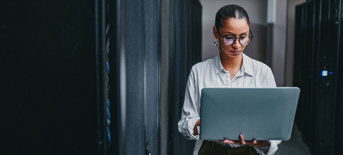 [Featured image] An IT support professional wearing glasses works on their laptop in a server room.