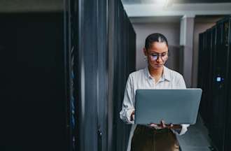 [Featured image] An IT support professional wearing glasses works on her laptop in a server room.