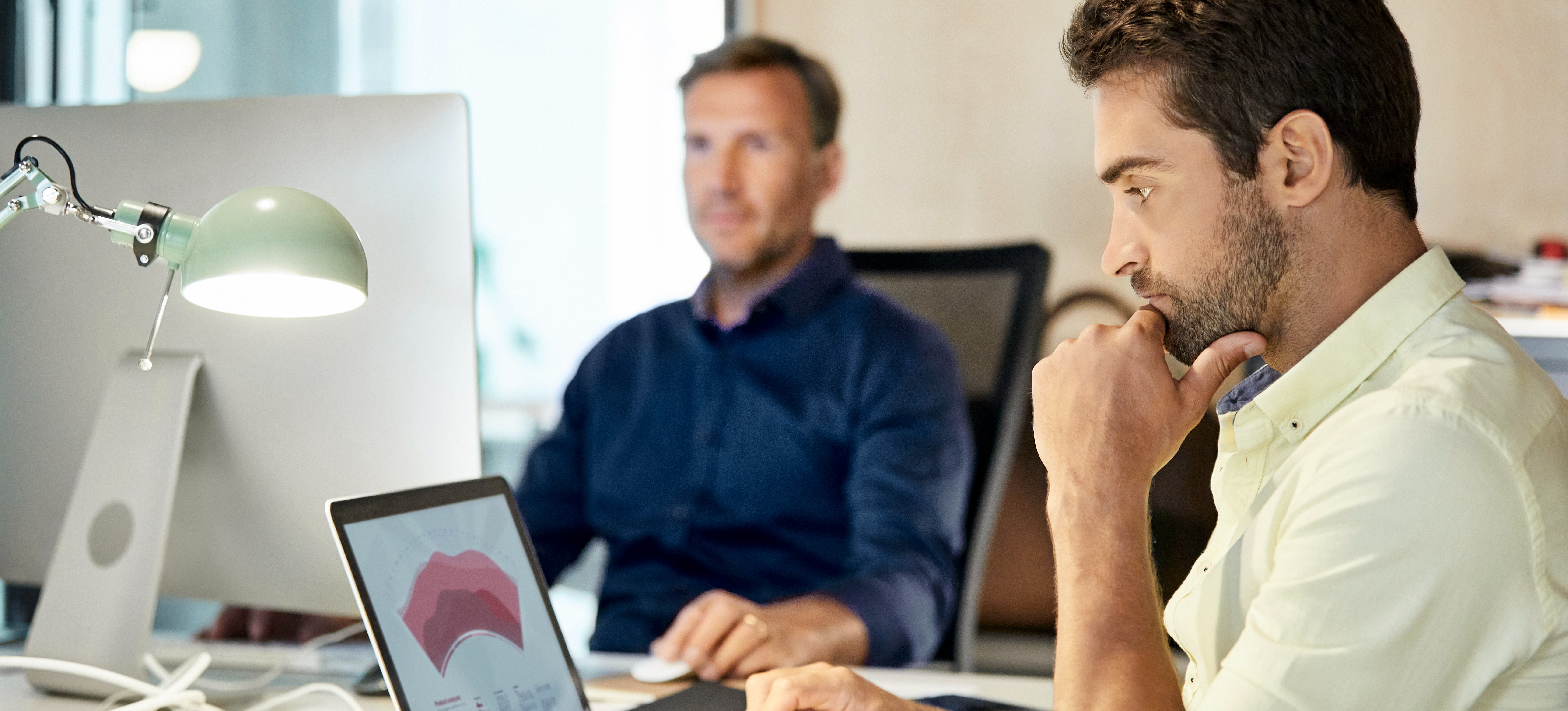 [Featured Image] Two colleagues sit at a desk looking at separate computers. Both of them are putting together their cloud computing projects before presenting to their supervisors.