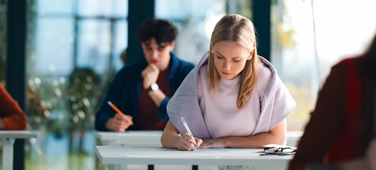 [Featured Image]: A learner uses ChatGPT for exam preparation while writing at a desk in a bright, modern classroom, while other learners work in the background.