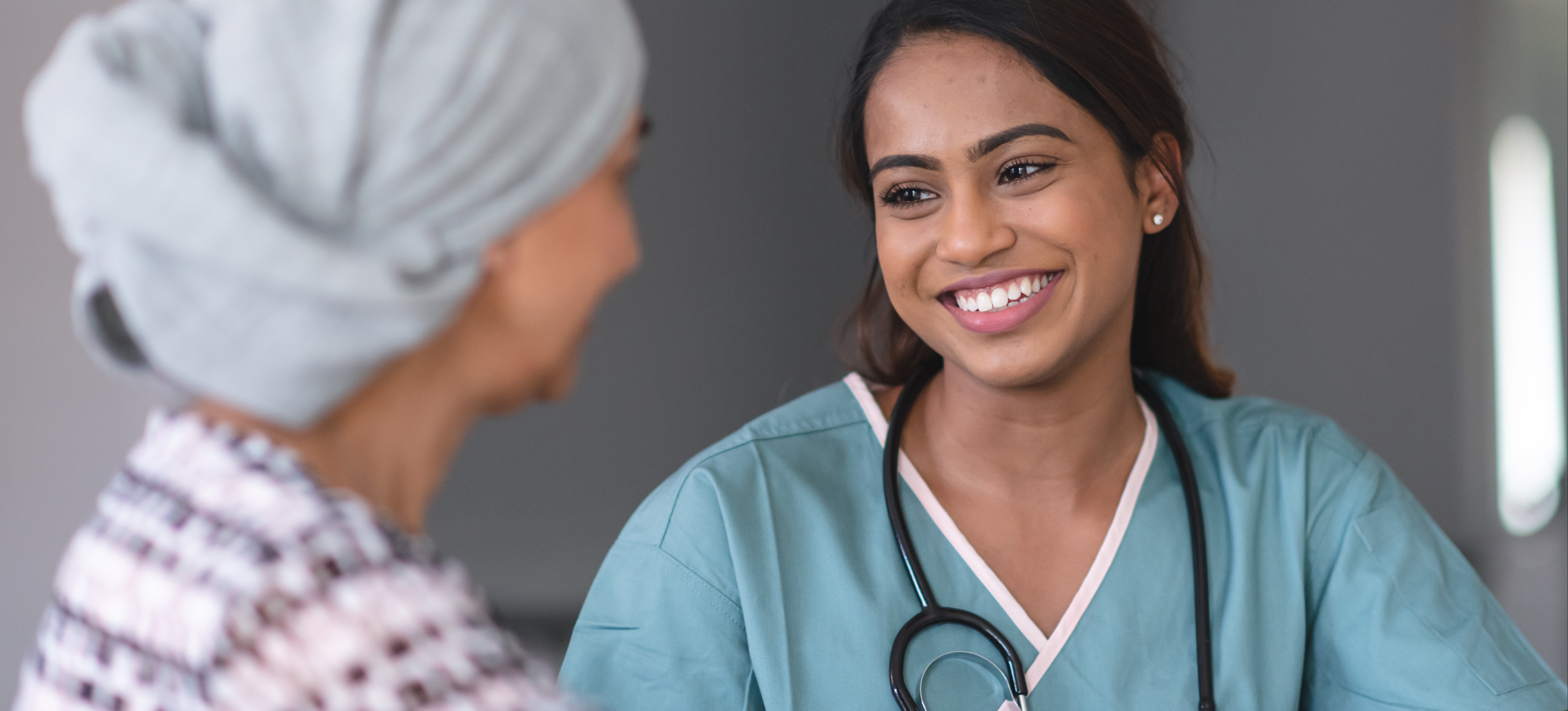 [Featured Image] An oncology nurse wearing light blue scrubs and a stethoscope around their neck speaks with her patient, who is wearing a black and white sweater and a head covering. 
