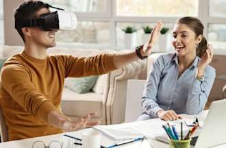 [Featured Image] Two extended reality (XR) developers sit in an office and experiment with a VR headset. 
