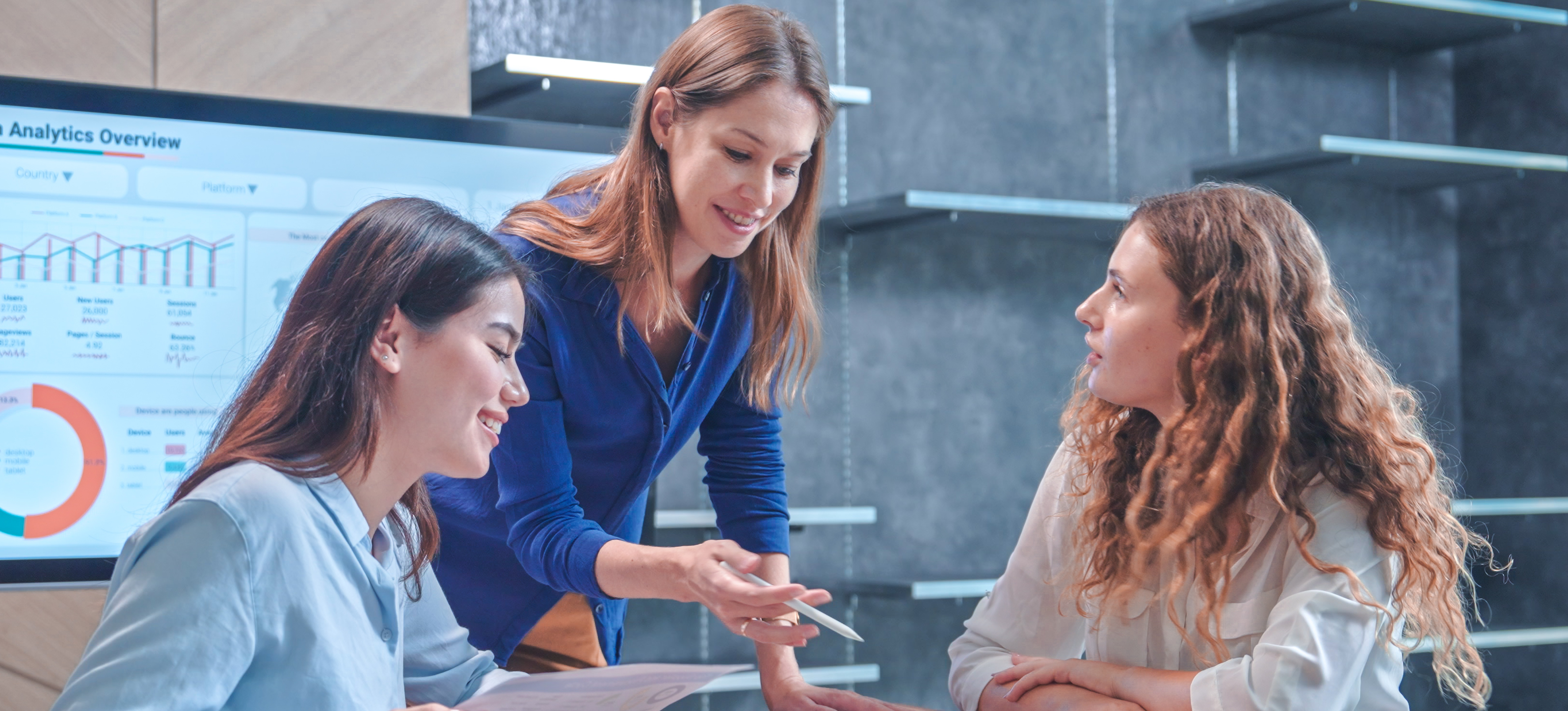 [Featured image] Three businesswomen brainstorm data analysis techniques in a modern office meeting room.
