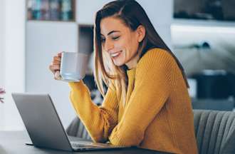 [Featured image] A project manager in a yellow sweater holds a mug and studies for the CAPM on a laptop at a desk with a small glass vase filled with pink flowers.