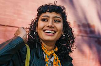 [Featured image] A young woman with curly black hair and big hoop earrings, wearing a black jacket and a color scarf, smiles at someone off-camera. 