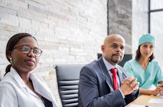 [Featured Image]:   A hospital administrator, wearing a suit and red tie, is sitting at his desk and consulting with two staff members, one wearing a white coat and the second wearing a green uniform.