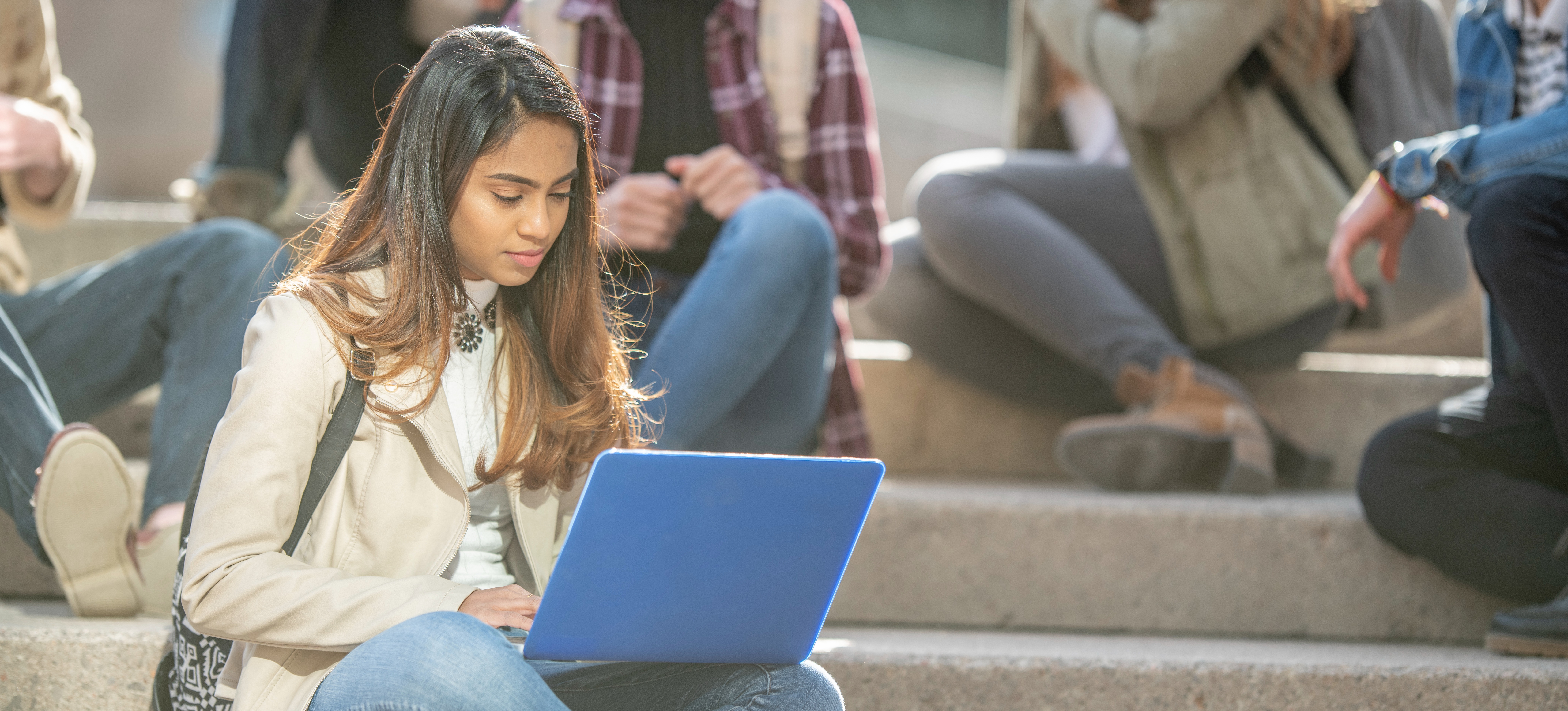 [Featured Image] Young university students sitting outside on stairs with a learner in the foreground researching master’s in data science course fees on a laptop.
