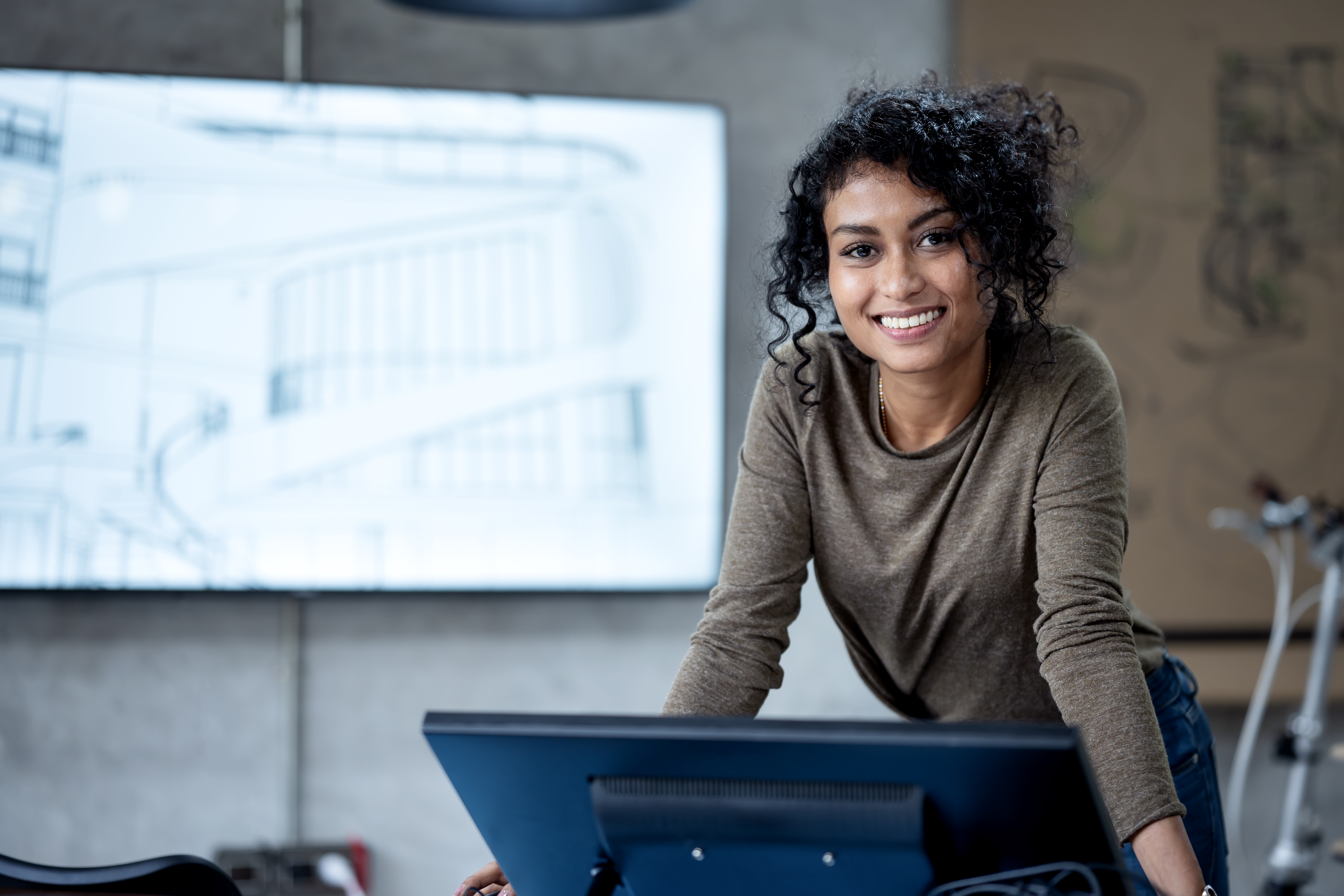 [Featured image] A woman in a brown shirt stands in front of a computer monitor smiling at the camera.