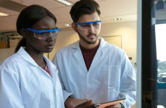 [Featured Image]: A female, with black hair, wearing a white uniform and blue goggles, and a male with black hair, wearing a white uniform and blue goggles. They are working in a lab, holding, writing and analyzing an experiment. 