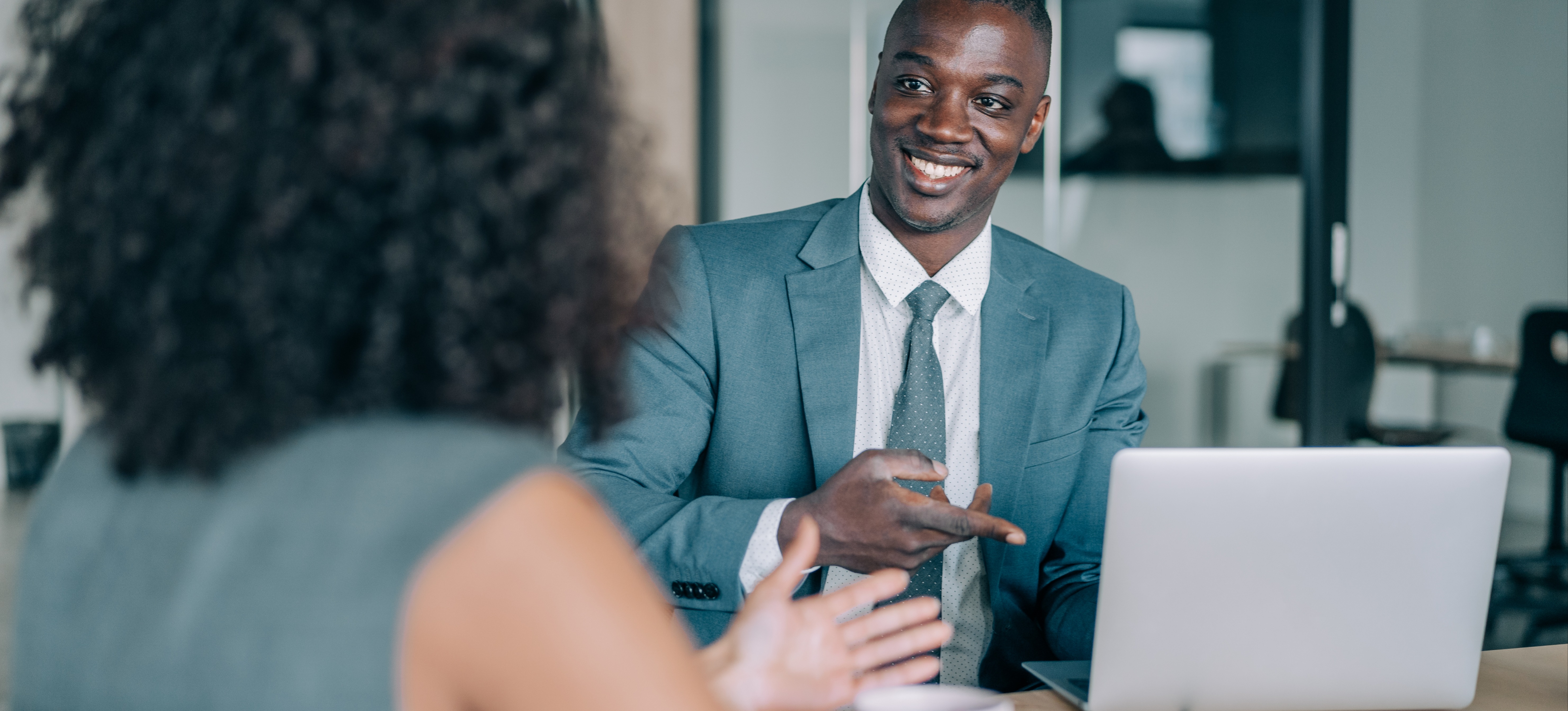 [Featured Image] A salesforce developer points at their laptop on a table during a meeting with a businesswoman.
