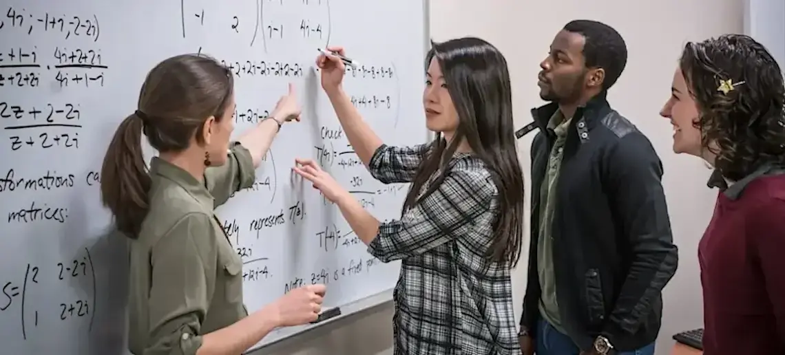 [Featured Image] A group of students stand in a classroom with an instructor teaching mathematics related to AI on a whiteboard.
