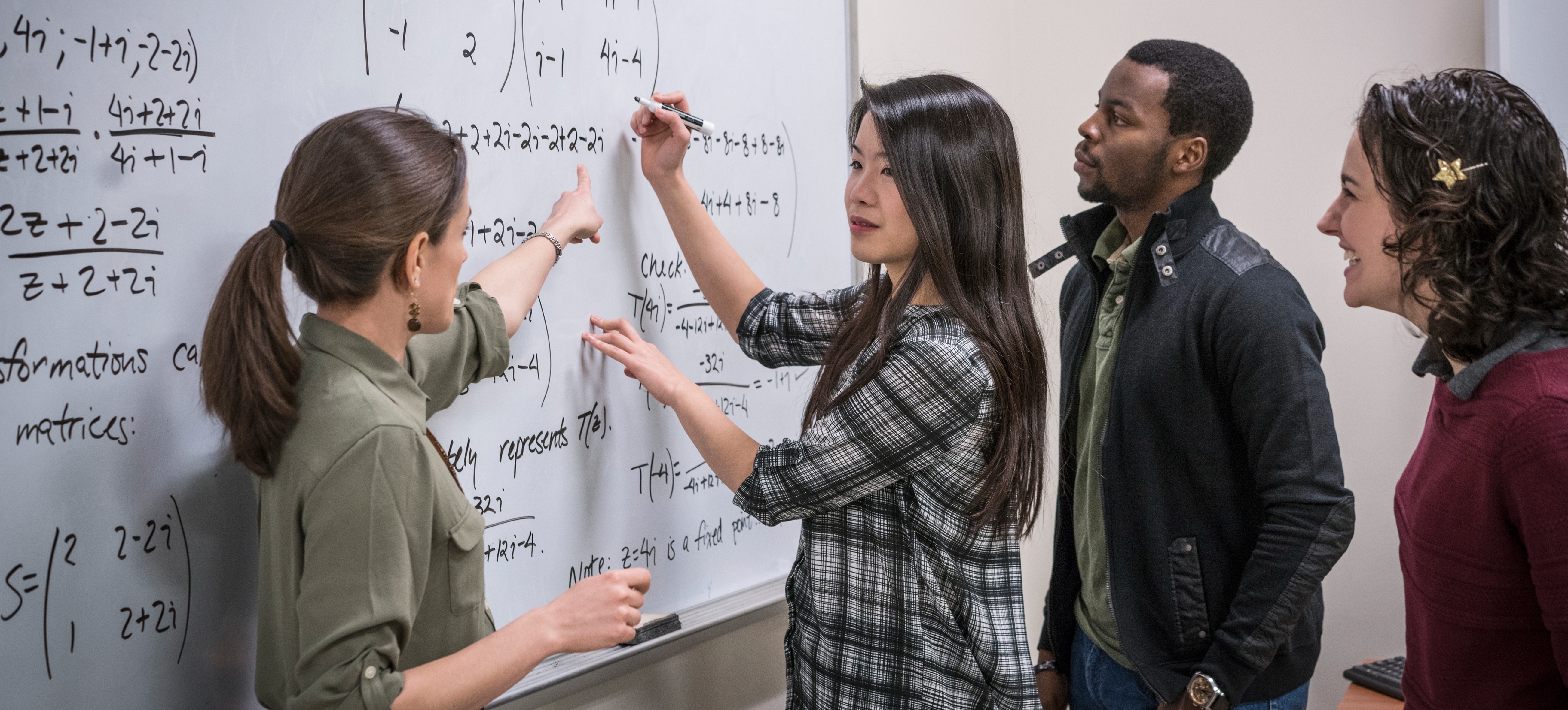 [Featured Image] A group of students stand in a classroom with an instructor teaching mathematics related to AI on a whiteboard.
